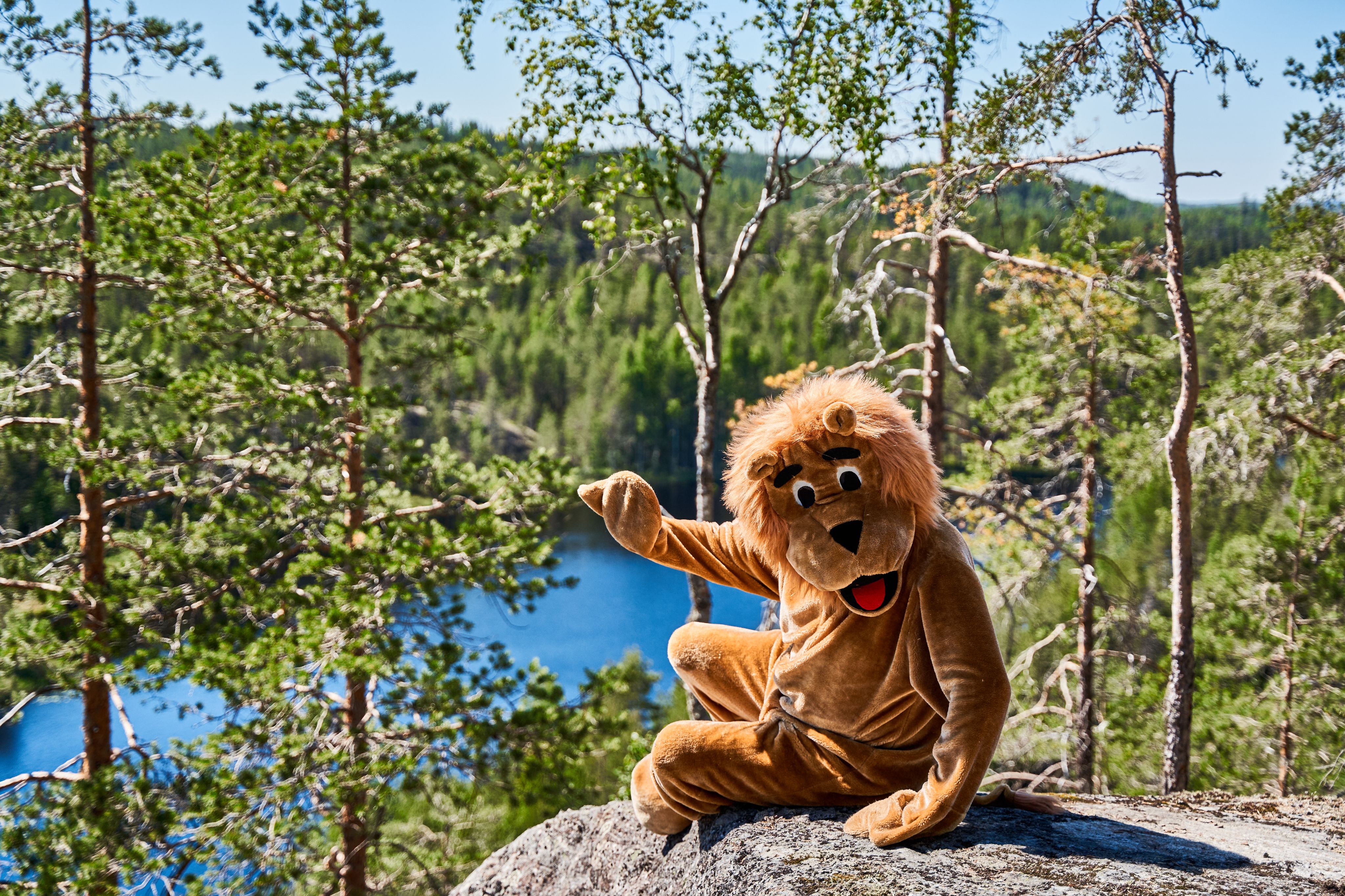 leijanaksi pukeutunut ihminen tervehtii kalliomaisemassa, taustalla metsää ja järvi